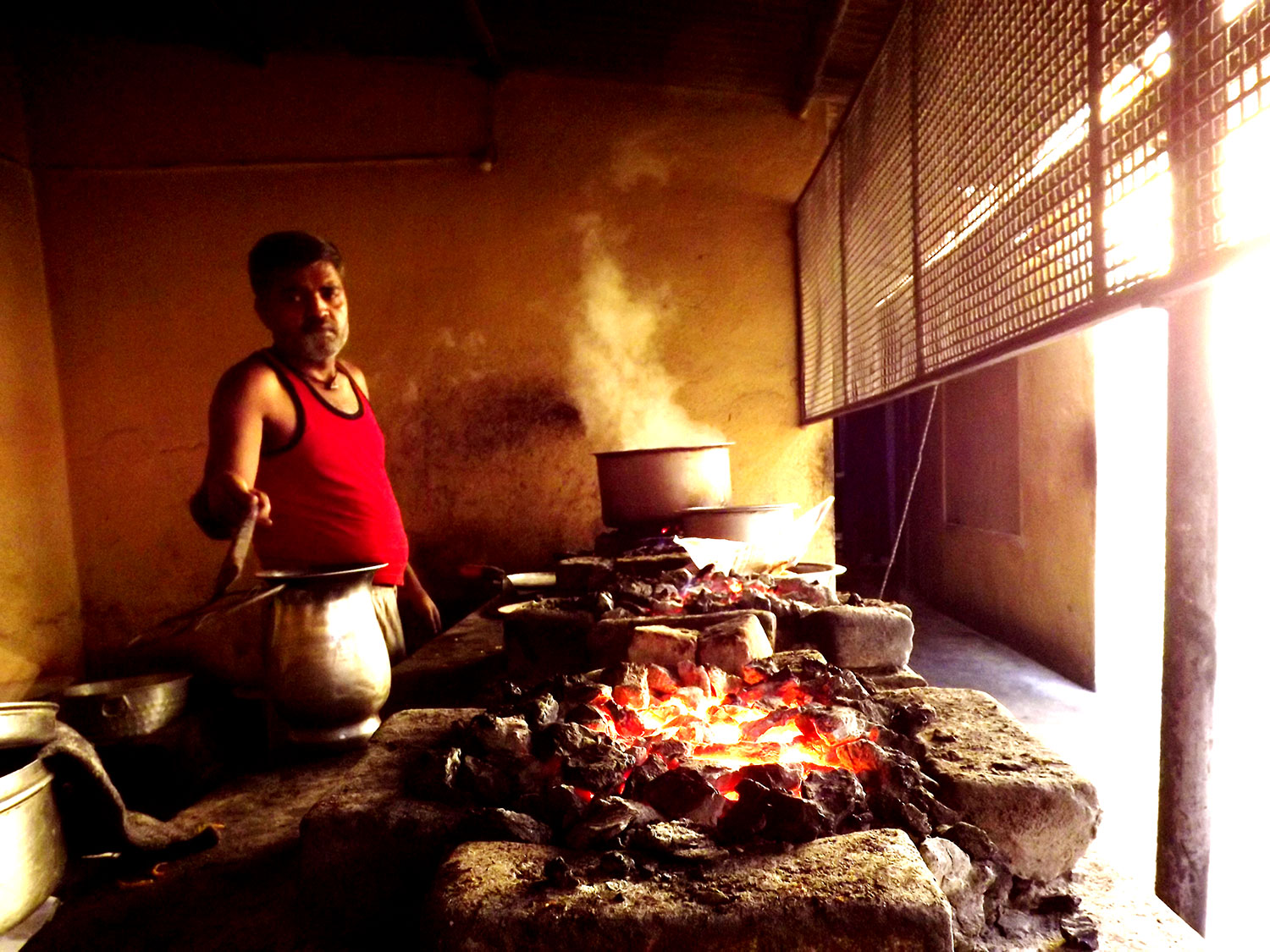 This photo shows a cook dressed in a red vest reaching out with a ladle next of a earthen clay oven with pits filled with smoldering charcoal brick in Ranchi, Jharkhand, India