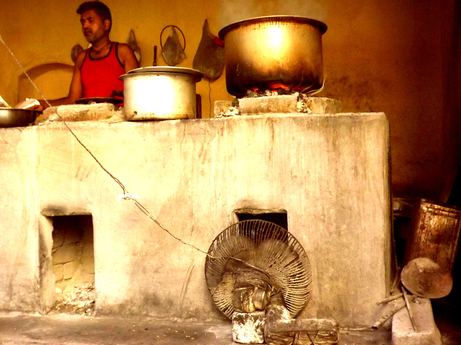 This photo show a section of a native clay oven from the front. Visible is a modified wall fan placed in front of a vent near the ground. On top of the clay oven are large bronze and metal containers.