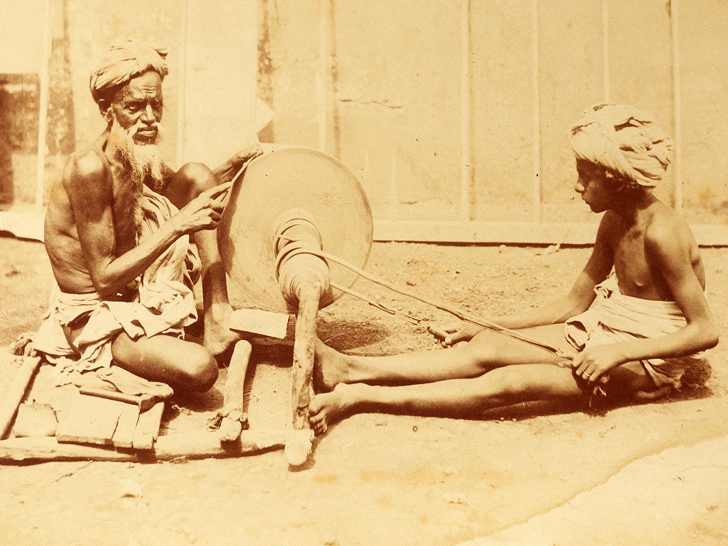 View of a scantily clad Mohammedan man sharpening a knife over a rotating whetstone along with a young boy, 1860