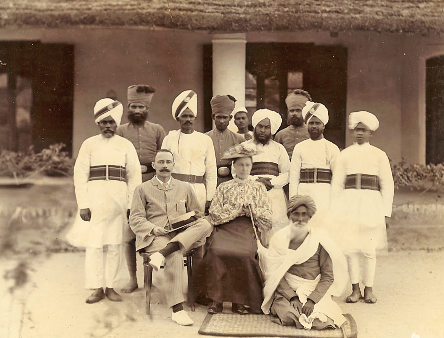 Image shows a vintage photo of a Euroepan couple amidst a collective of native policemen in British India, 1900.