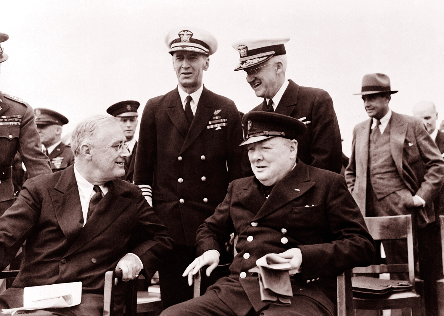 Image shows a vinate photo of U.S President Roosevelt and British Prime MInister Winston Churchill sitting side by side on the deck of the HMS Prince of Wales.