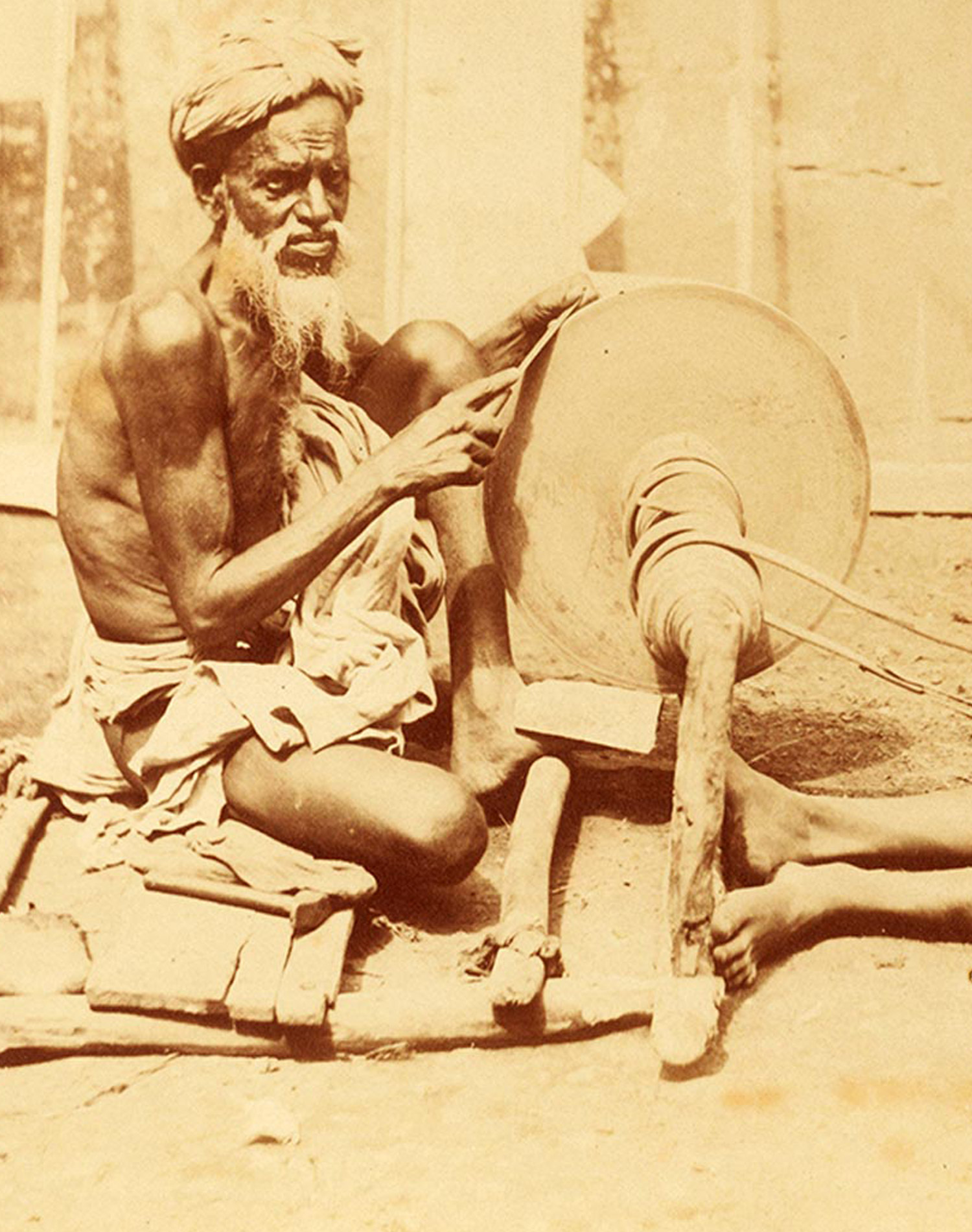 A vintage photo showing a scantily clad Mohammedan man sharpening a knife over a rotating whetstone along with a young boy, 1860